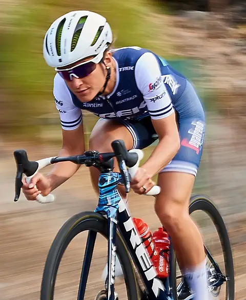 A female cyclist wearing a white helmet, sunglasses, and a blue, white, and red Trek-Segafredo team cycling kit is shown mid-pedal on a road bike