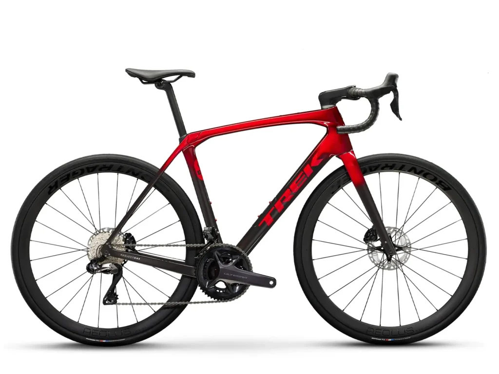 A side view of a Trek road bike in a red and black color scheme. It has narrow tires and a carbon frame