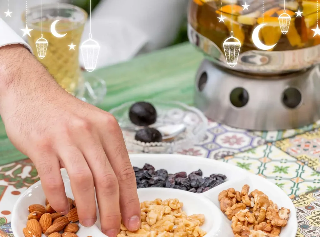 A person's hand reaches for nuts and dried fruits on a plate, with a pot and dates nearby. The scene is decorated with Ramadan-themed elements.