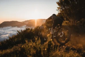 Cyclist climbs a rocky mountain trail at sunset, dust rising from the wheels, with golden backlight highlighting the motion.
