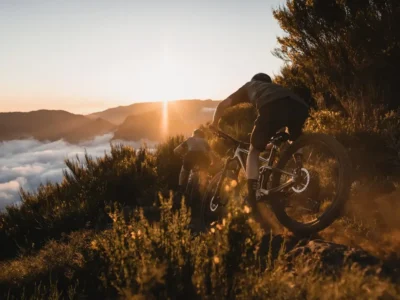 Cyclist climbs a rocky mountain trail at sunset, dust rising from the wheels, with golden backlight highlighting the motion.
