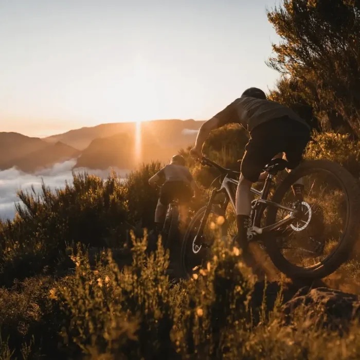 Cyclist climbs a rocky mountain trail at sunset, dust rising from the wheels, with golden backlight highlighting the motion.