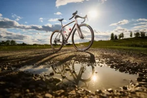 The image shows a Trek bicycle, likely a gravel or road bike, standing on a dirt path next to a puddle. The bright sun in the background is reflected on the water's surface, mirroring the bike.