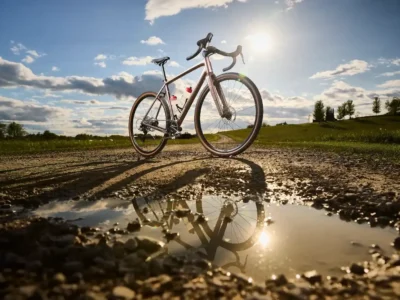 The image shows a Trek bicycle, likely a gravel or road bike, standing on a dirt path next to a puddle. The bright sun in the background is reflected on the water's surface, mirroring the bike.