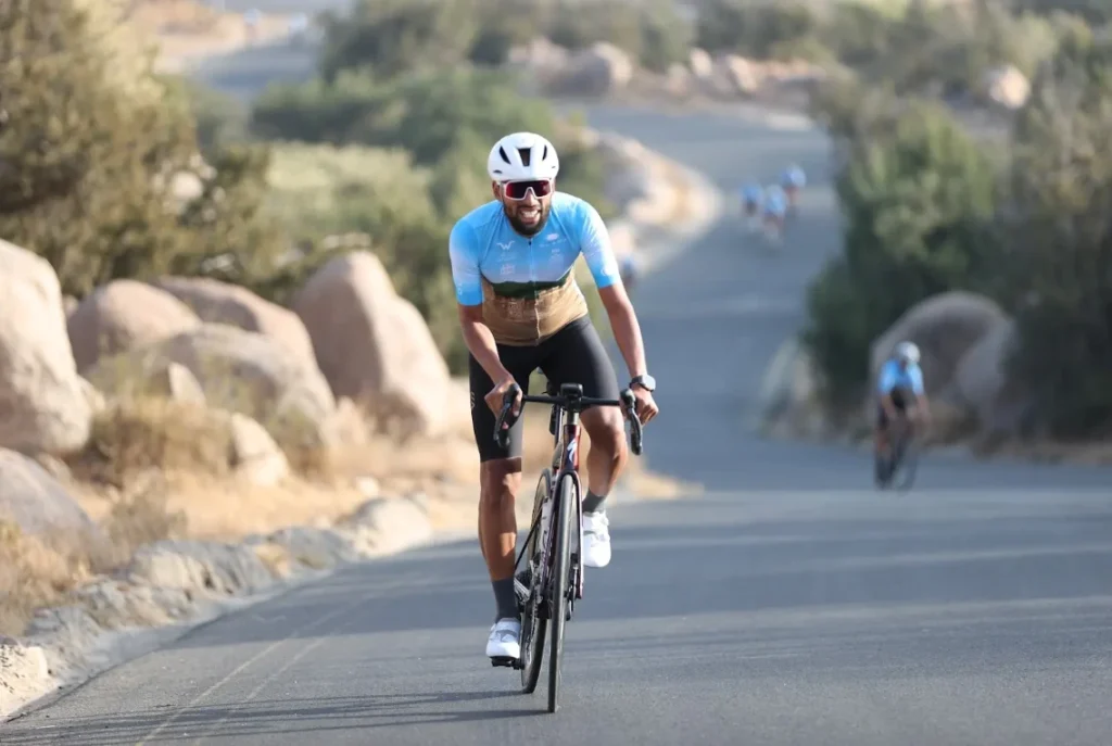 Cyclist in blue jersey and white helmet riding uphill on a paved road surrounded by rocks and trees on a sunny day.