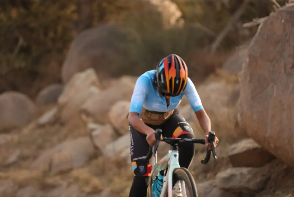 Cyclist wearing light blue jersey and orange helmet, riding on a rocky, desert-like path with focused posture and determination.