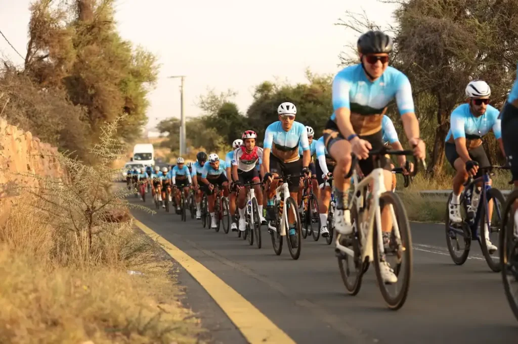A group of cyclists riding on a clear road surrounded by trees, wearing blue jerseys and helmets, focused and moving forward.