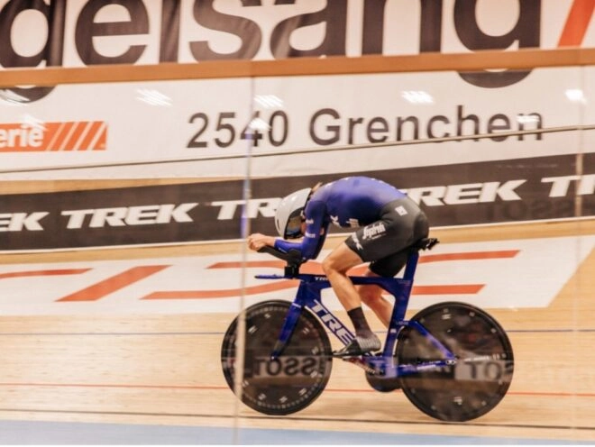 Low-angle shot of an indoor cycling race, featuring a professional cyclist on a blue bike on the track, wearing a white helmet and dark blue kit.