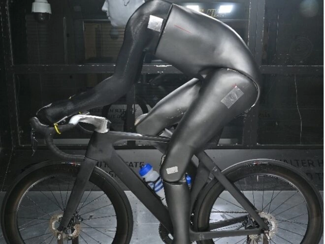 Manny, a black-suited robot on a black bike in a wind tunnel, mimics a rider to test aerodynamics and precise adjustments.